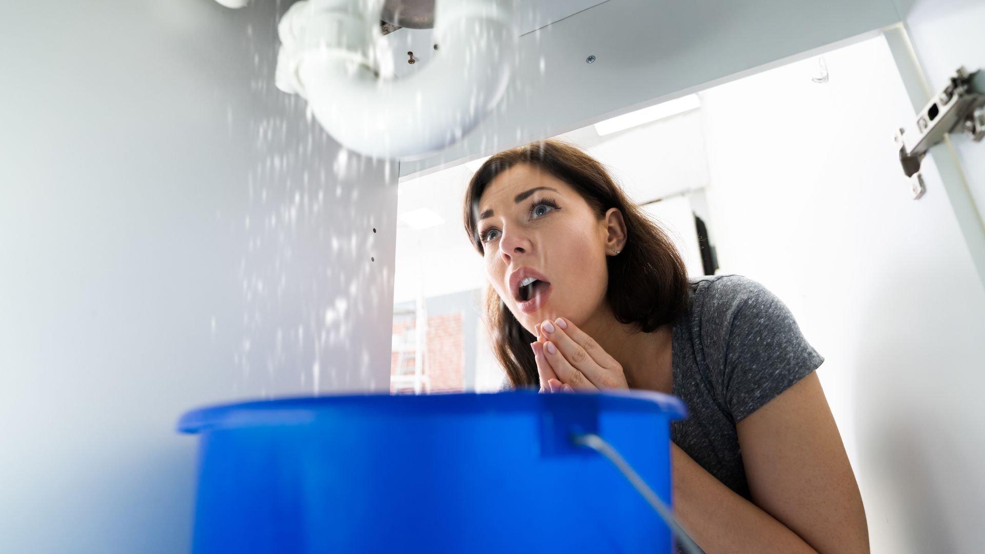 Shocked woman looking at water leaking from ceiling into blue bucket