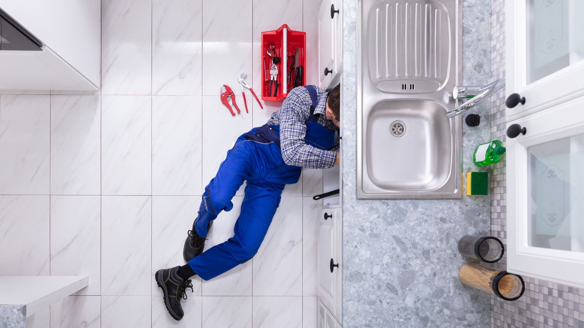 Plumber in blue overalls fixing kitchen sink from overhead view